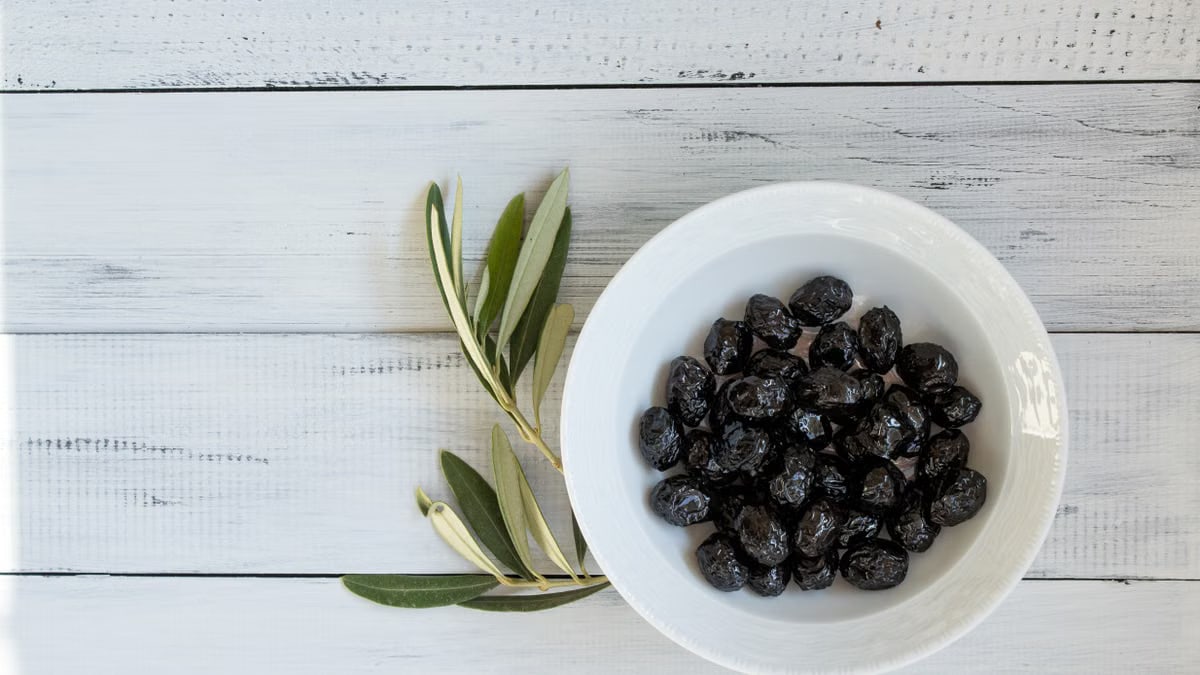 A bowl of black olives on a rustic white wooden surface with olive leaves. Perfect for culinary themes.